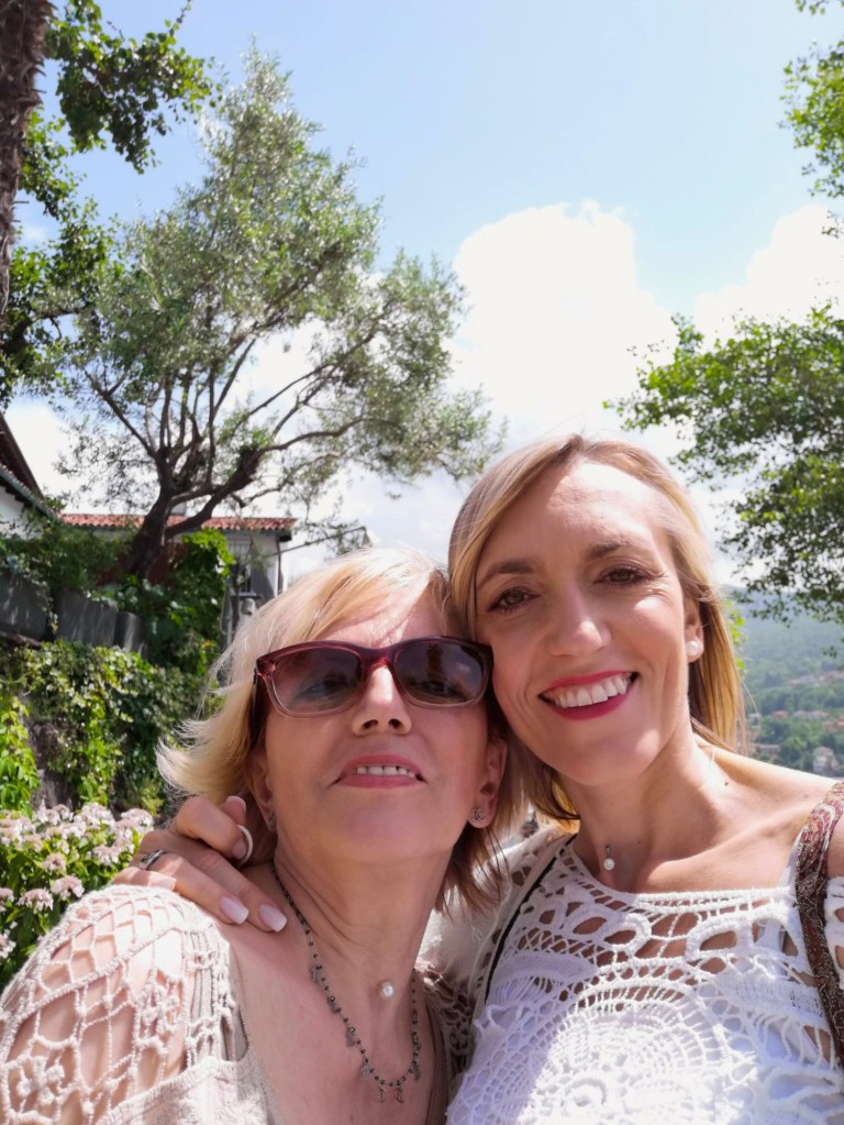 Due donne sorridenti in primo piano, con occhialini da sole, in un ambiente verdeggiante e soleggiato.