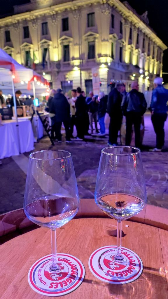 Due bicchieri di vino su un tavolo attorno a una festa serale all'aperto, con persone e bancarelle illuminate sullo sfondo.