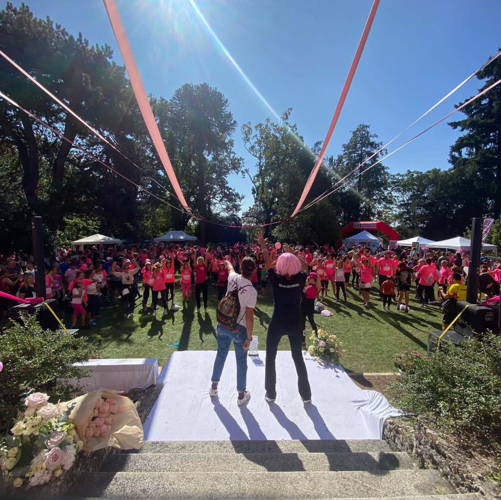 Persone in abiti rosa partecipano a un evento all'aperto, con due persone che si trovano sul palco di fronte a un pubblico numeroso.