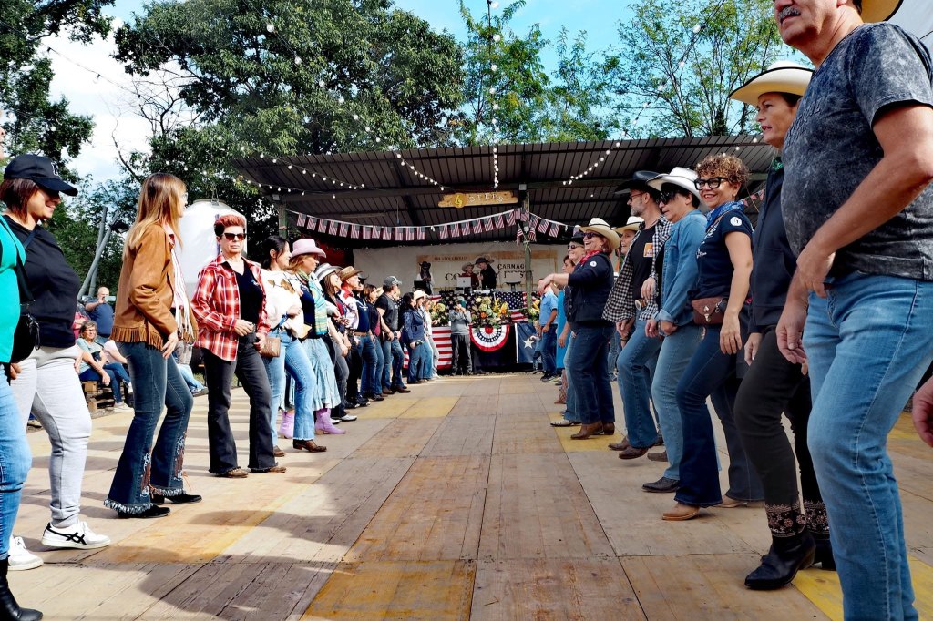 Gruppo di persone che partecipano a una danza, indossando abbigliamento da cowboy, su un palco decorato con bandiere. Sullo sfondo si intravede un pubblico e un'area di intrattenimento.