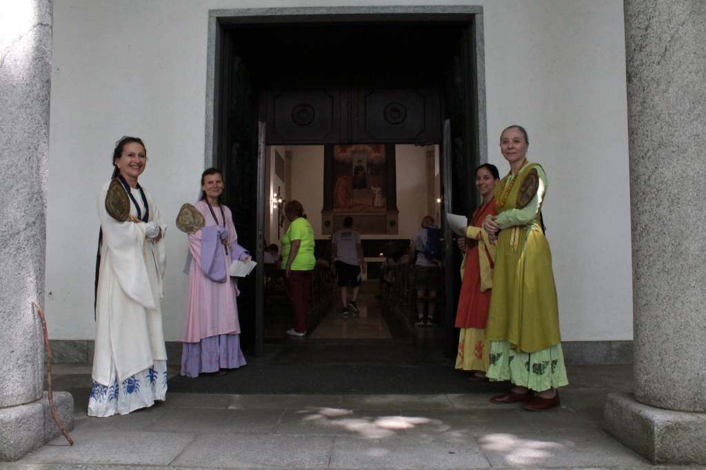 Donne in abiti tradizionali sorridono all'ingresso di un edificio, con un dipinto visibile all'interno.
