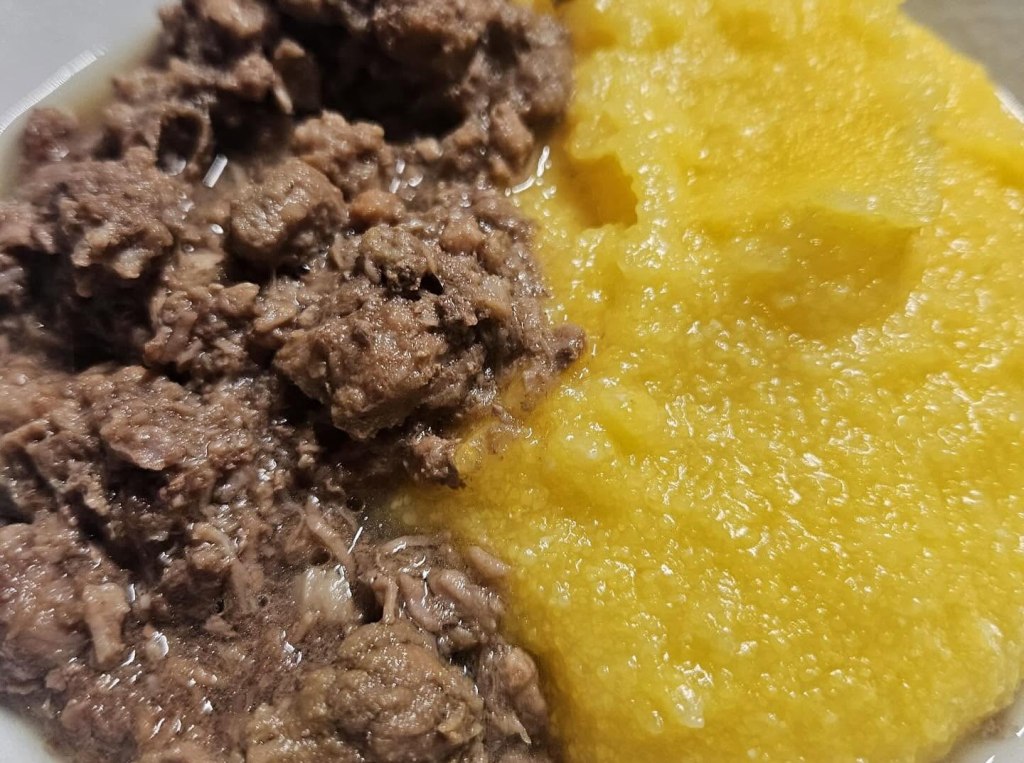 Piatto di carne tritata aromatizzata servito accanto a una porzione di polenta gialla cremosa.