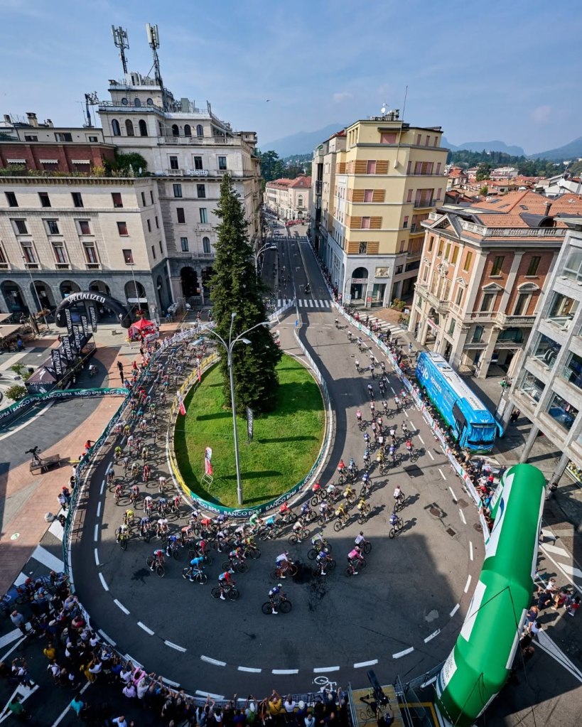 Una panoramica di una corsa ciclistica in una città, con ciclisti che curveggiano attorno a una rotonda. Edifici storici e moderni circondano la scena, mentre una folla osserva l'evento.