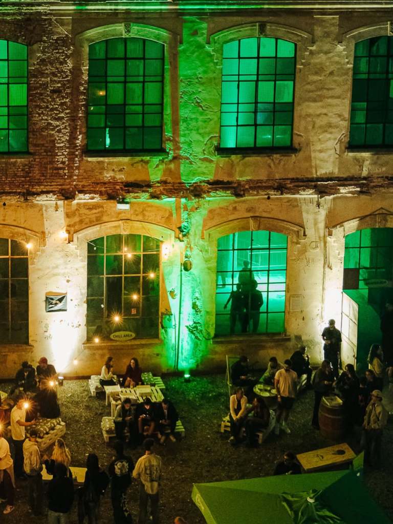 Una vista notturna di un evento all'aperto in un'area industriale illuminata di verde, con persone che si godono la serata intorno a tavoli e sedie.