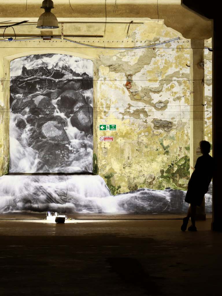 Murale di un fiume che scorre su una parete danneggiata, con un'illuminazione soffusa e una figura umana in siluetta sul lato destro.