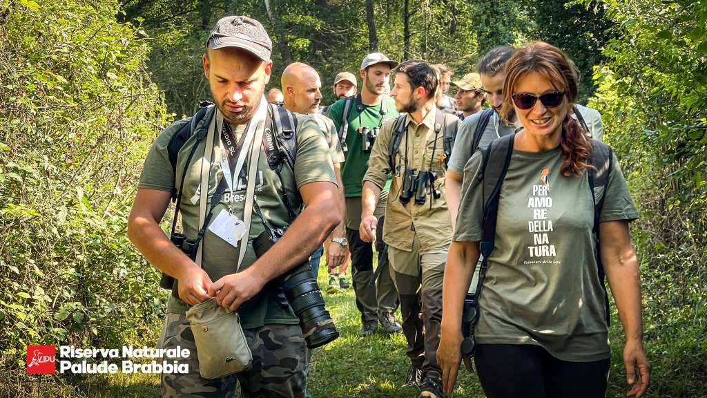Gruppo di persone in cammino nella Riserva Naturale Palude Brabbia, vestiti in abbigliamento per escursioni, alcuni con binocoli e fotocamere.