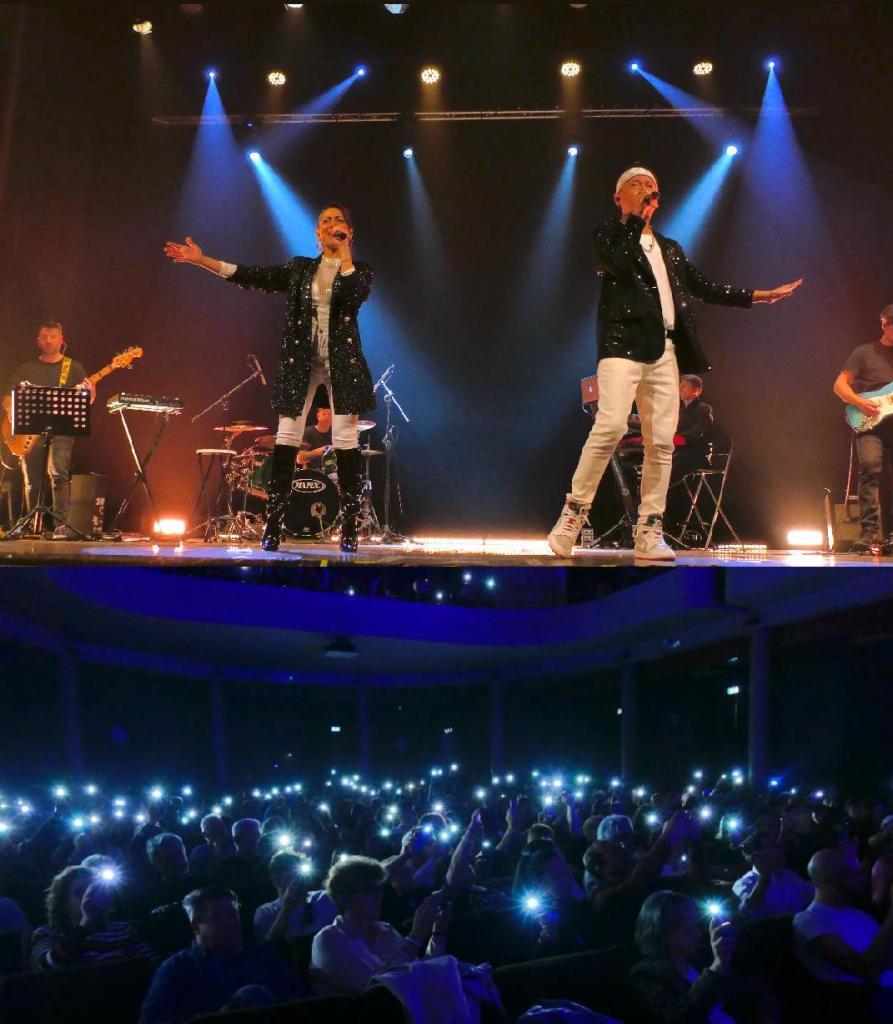Artisti sul palco durante un concerto, con luci colorate e band che suona dietro di loro.