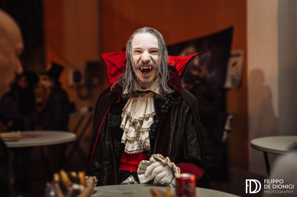 Un uomo vestito da vampiro con un mantello rosso e nero, capelli lunghi e bianchi, sorridente mentre tiene un oggetto in mano.