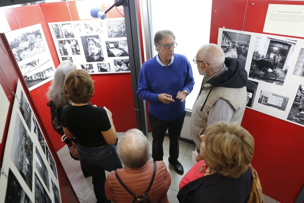 Un gruppo di persone discute davanti a una mostra fotografica che presenta immagini in bianco e nero su un muro rosso.