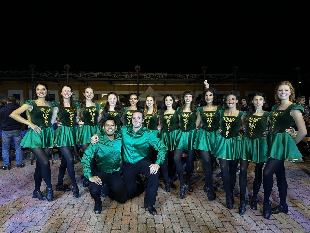 Un gruppo di ballerini in costumi verdi, con abiti abbelliti da simboli dorati, posano insieme in un evento all'aperto di notte.