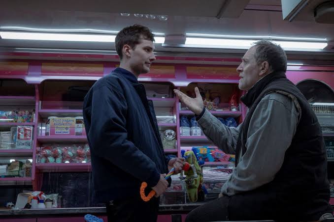 Due uomini stanno parlando all'interno di un negozio colorato, circondati da giocattoli e articoli vari. Uno tiene un giocattolo nella mano.