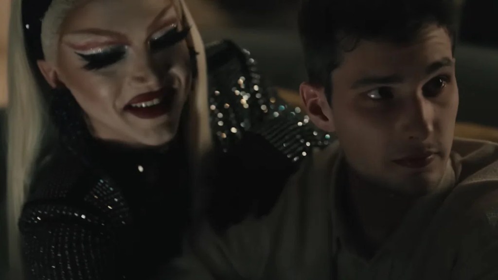 Una persona truccata con un trucco teatrale e capelli lunghi, vestita in modo glamour, sorride mentre si avvicina a un giovane che sembra sorpreso o sconcertato.