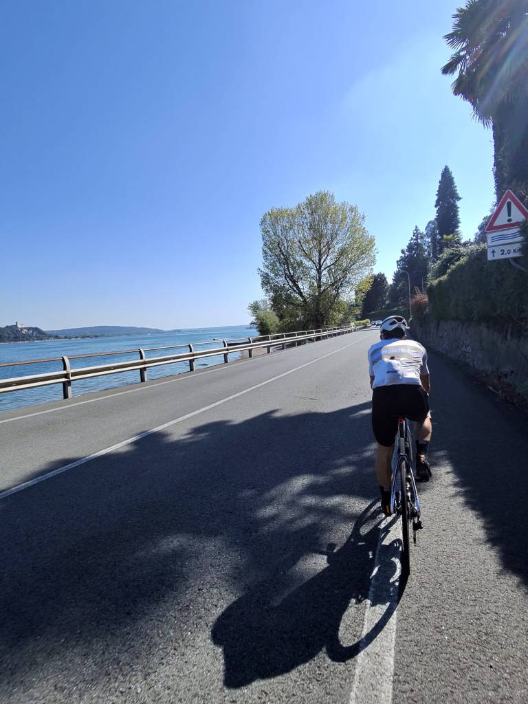 Un ciclista su una strada asfaltata lungo un lago, con alberi e una strada di legno sulla sinistra. Il cielo è sereno e blu.