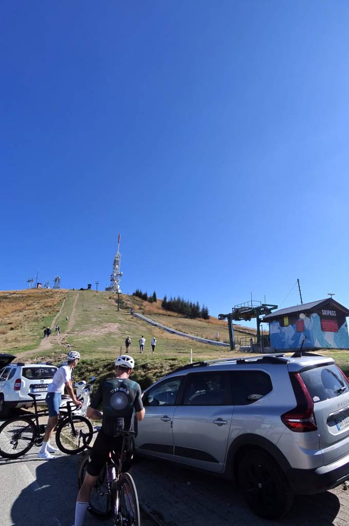 Vista di una montagna con ciclisti e pedoni, parcheggiati alcuni veicoli, sotto un cielo blu sereno.