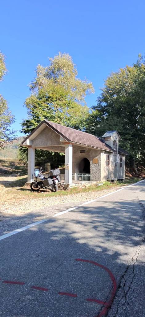 Una piccola cappella accanto a una strada, con un motociclo parcheggiato davanti e alberi sullo sfondo.