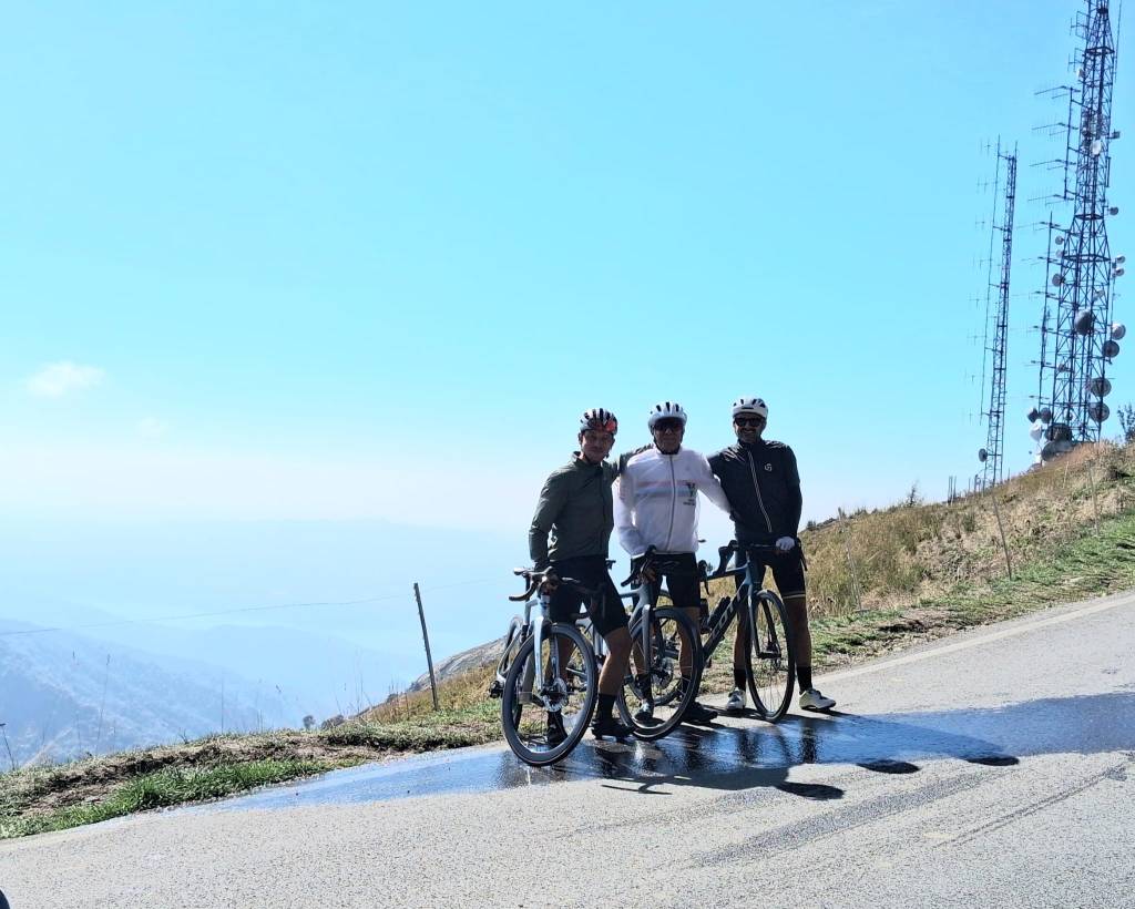 Tre ciclisti in posa su una strada di montagna, con torri di comunicazione sullo sfondo e cielo azzurro.